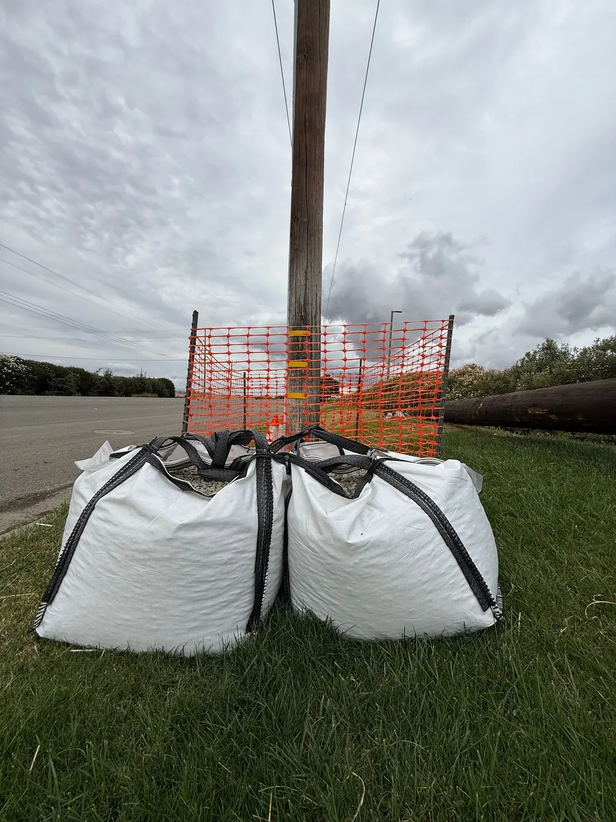 Filled bulk bags anchoring a utility pole base behind orange safety fencing on a roadside job